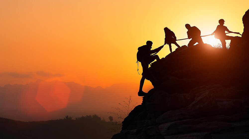 Silhouette des Kletterteams, das sich gegenseitig beim Klettern bei einem Sonnenuntergang hilft.