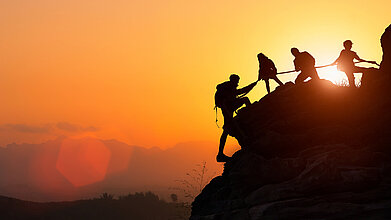 Silhouette des Kletterteams, das sich gegenseitig beim Klettern bei einem Sonnenuntergang hilft. Der Begriff der Beihilfe.