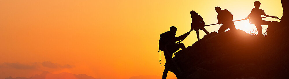 Silhouette des Kletterteams, das sich gegenseitig beim Klettern bei einem Sonnenuntergang hilft. Der Begriff der Beihilfe.