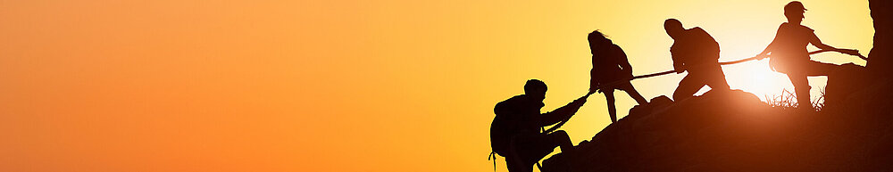 Silhouette des Kletterteams, das sich gegenseitig beim Klettern bei einem Sonnenuntergang hilft. Der Begriff der Beihilfe.