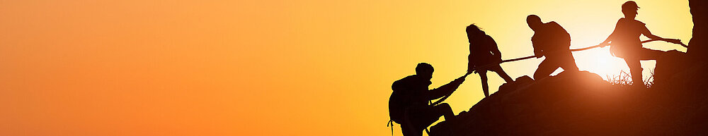 ©iStock\sorn340 Silhouette des Kletterteams, das sich gegenseitig beim Klettern bei einem Sonnenuntergang hilft. Der Begriff der Beihilfe.