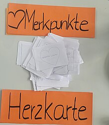 Fotografie: Herzkarten; © Anna Wenzl; Begriffe auf organgem Karton: Herzsymbol, Merkpunkte; Herzkarte; in der Mitte ein weißer Papierstapel, oberstes Blatt: "denken wagen"
